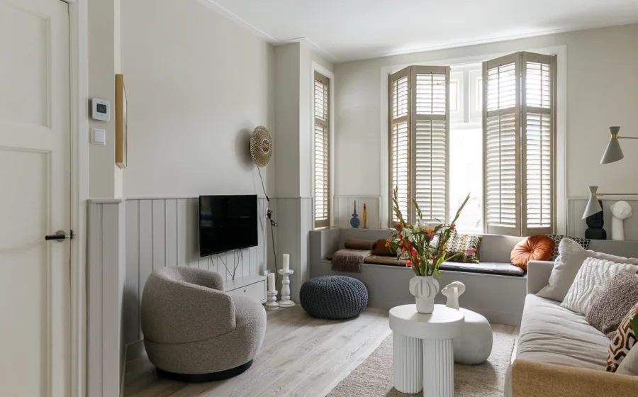 Woonkamer met glas-in-loodramen met houten shutters en muur met lichtgrijze houten lambrisering.