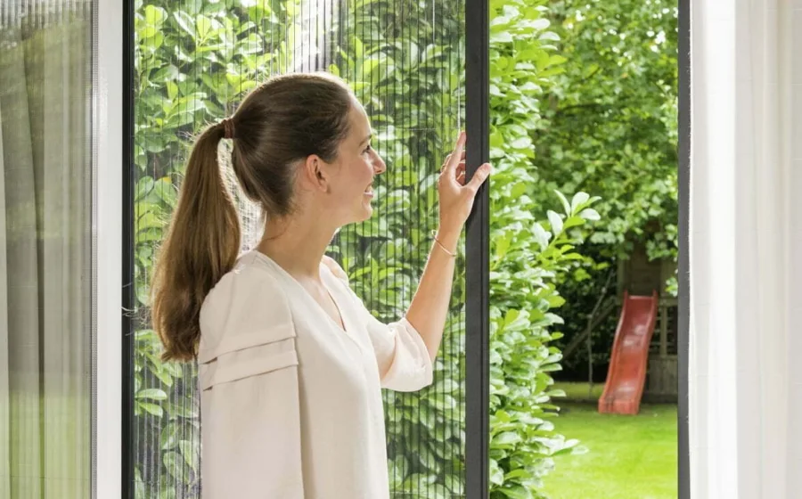 Vrouw schuift een plissé hor van Unilux open voor een geopend draaikiepraam, met zicht op een groene tuin