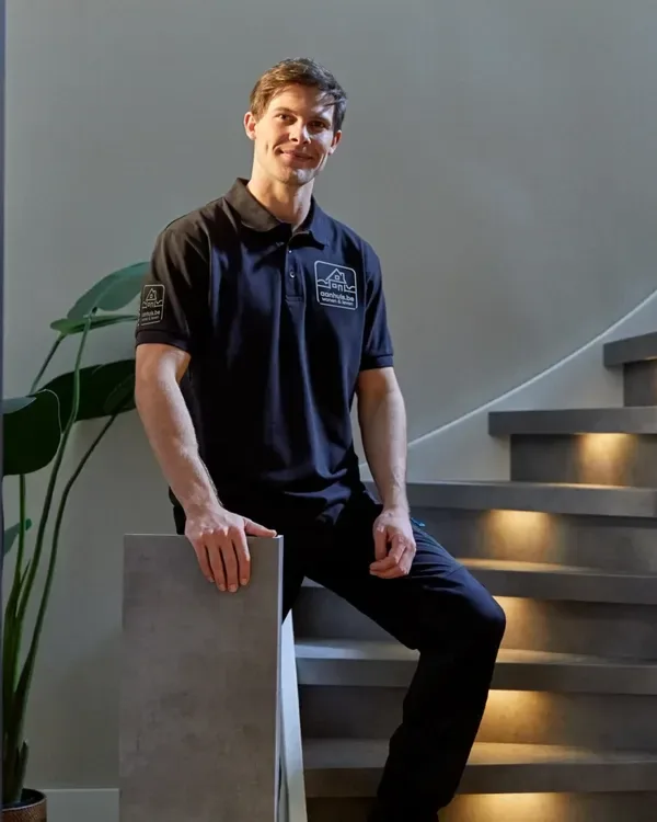 Medewerker van aanhuis.be in een donkerblauwe polo met logo zit op een betonlook trap met verlichte treden